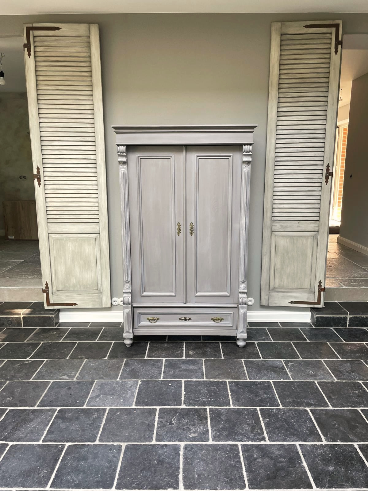 Taubengrau gefasster Schrank zwischen zwei Fensterläden
